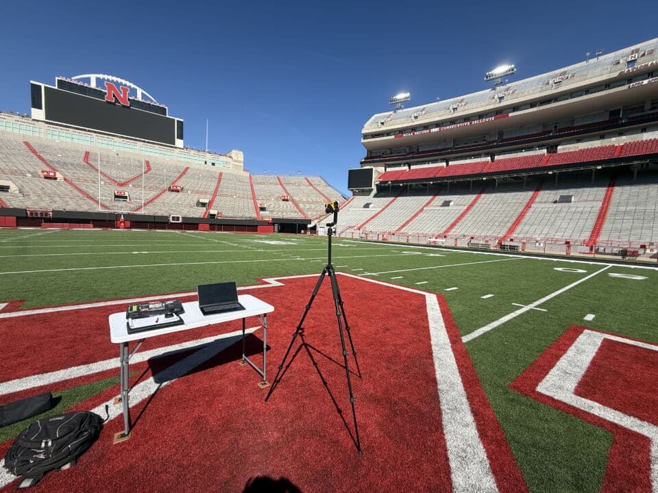 An Audio Engineer's TruPulse 360i, stands mounted onto a tripod and next to some gear, in the middle of Nebraska's Memorial Stadium